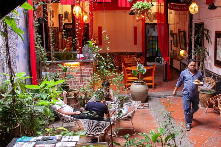 Vredig binnenhof van een Maleisisch restaurant met groene planten, roodkleurige decoratie, gezellige verlichting en een gast die genieten van een maaltijd, typische sfeer van veelzijdig Maleisië.