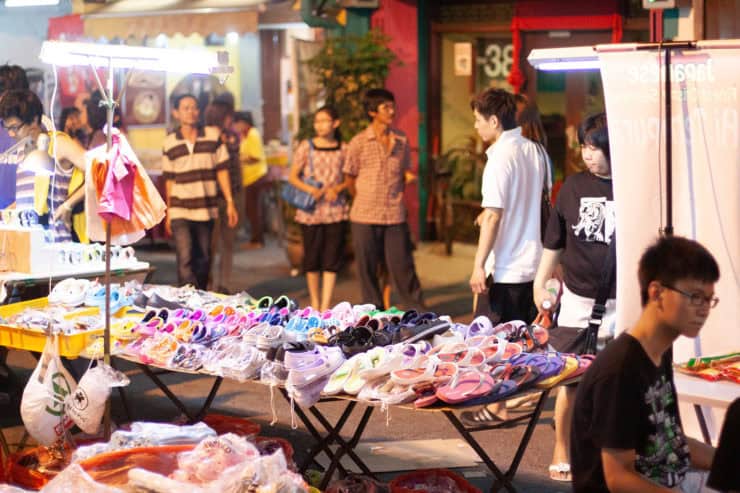 Kleurrijke zomersandalen en kindersandalen te koop op een markt in Maleisië, met een levendige sfeer en diverse kopers die genieten van lokale straatverkoop.